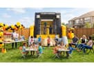 A large "Skid Loader" construction-themed inflatable bounce house and slide set up for a backyard birthday party. Children are shown playing inside the inflatable while others eat at child-sized tables. The event features a popcorn and cotton candy statio