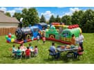 large, colorful train-themed inflatable obstacle course and bounce house setup for a backyard birthday party in Houston. The scene features children playing in the inflatable, a concession station with popcorn and cotton candy, and families seated at part